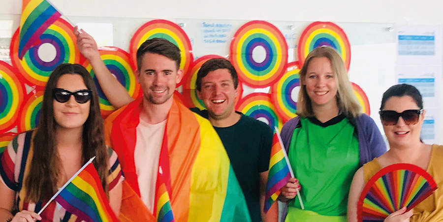 Un groupe de cinq personnes célébrant avec des drapeaux arc-en-ciel et des couleurs dans un cadre coloré, montrant fierté et diversité.