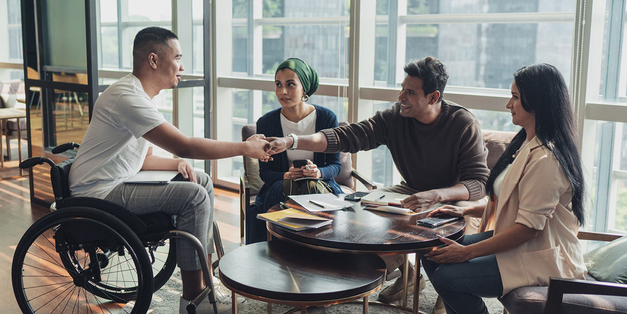 Un groupe de personnes s'assoit à une table et interagit. Des blocs-notes et des stylos sont posés sur la table. Deux hommes se serrent la main de l'autre côté de la table, l'un d'eux étant assis dans un fauteuil roulant. 