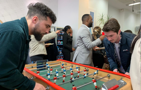 Un groupe de personnes socialisant lors d'un événement de bureau, avec deux hommes jouant au babyfoot au premier plan tandis que d'autres discutent et dégustent des rafraîchissements en arrière-plan.