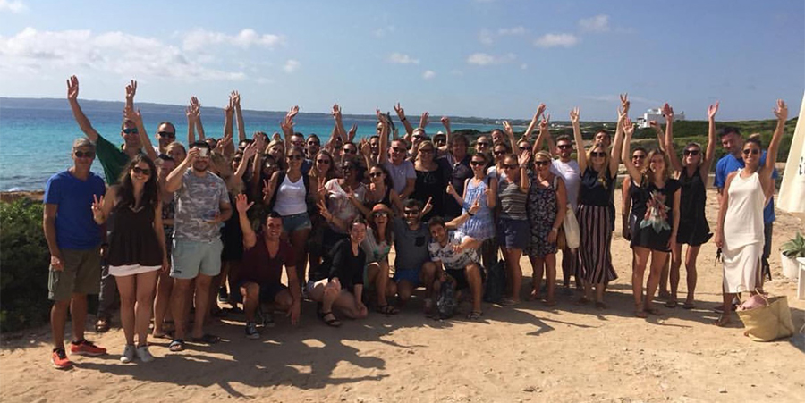 Un grand groupe de personnes posant joyeusement sur une plage de sable avec l'océan en arrière-plan sous un ciel bleu clair.