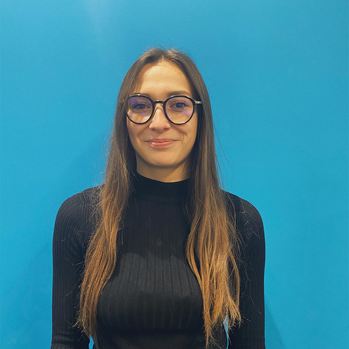  Femme aux cheveux longs et aux lunettes debout sur un fond bleu portant un haut noir.